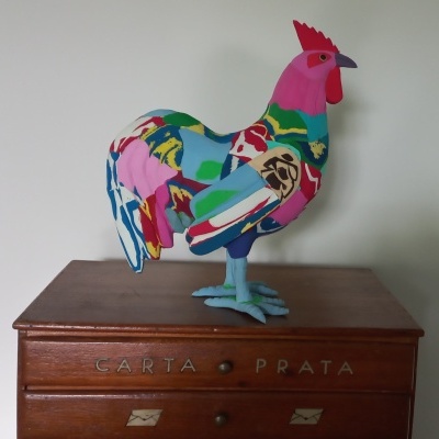 Escultura de galo em tecido multicolor sobre cómoda de madeira com texto