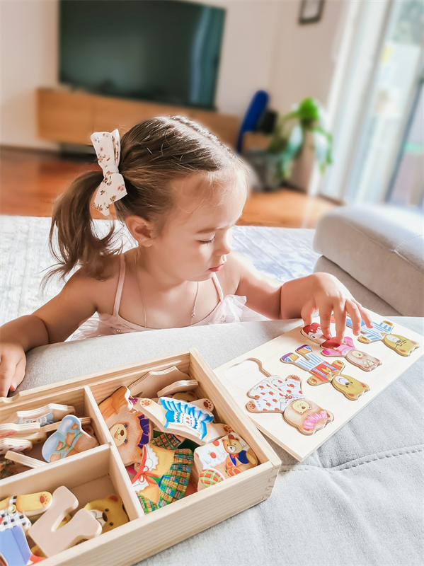 Criança a jogar com puzzle de madeira colorido em formato de bonecos, com caixa organizadora, numa sala de estar.