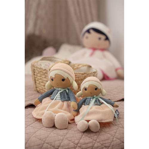 Dois bonecos de tecido com roupa cor de pêssego e cinzento sentados numa cama com colcha cor de rosa e um cesto ao fundo