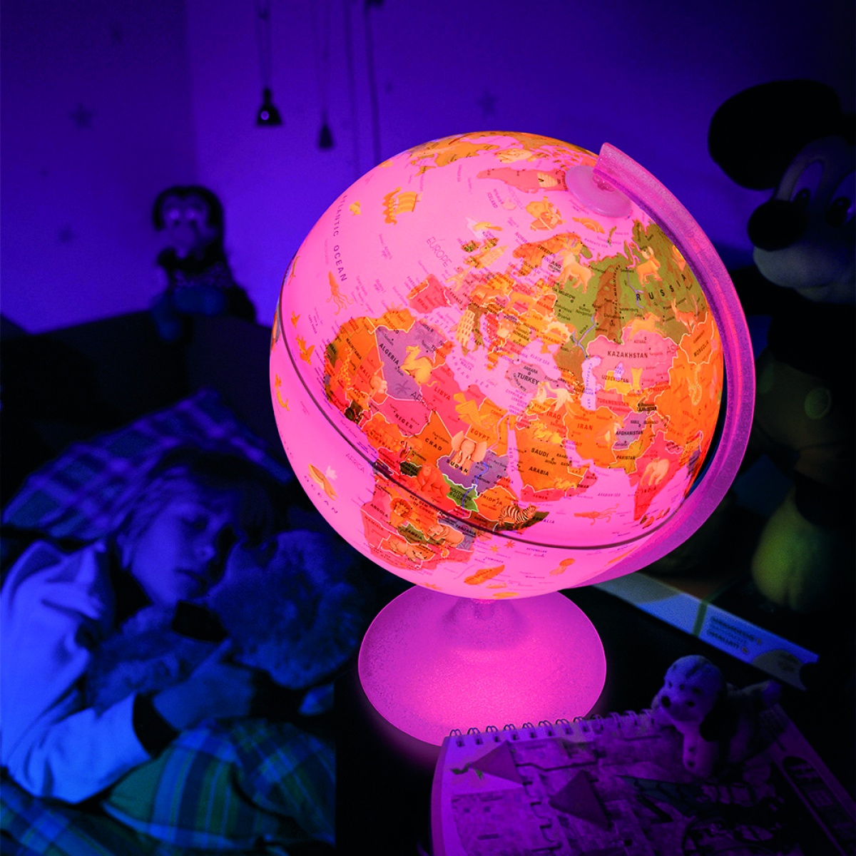 Globo terrestre iluminado cor-de-rosa numa mesa, com criança a dormir e brinquedos ao fundo
