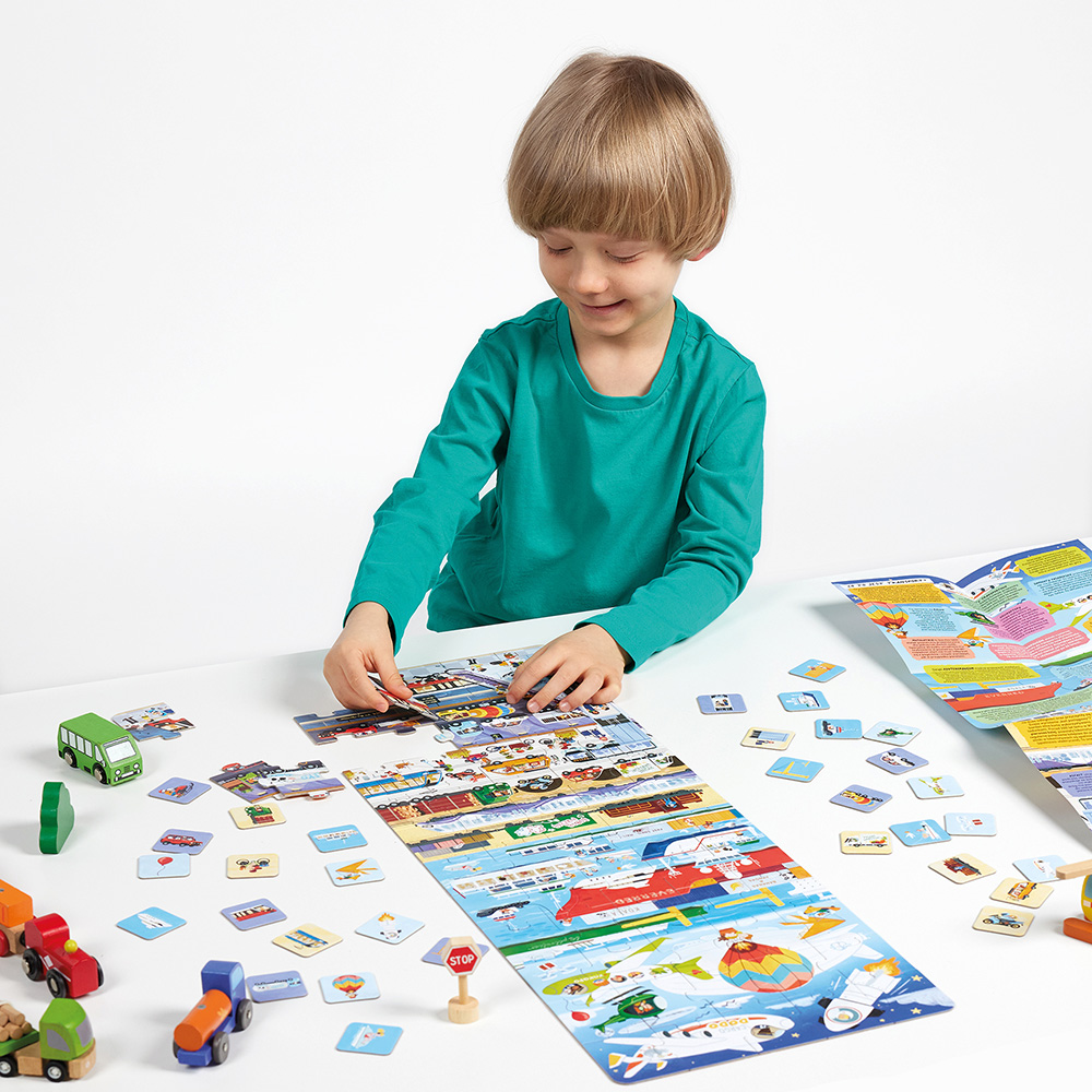 Criança a brincar com jogo educativo de peças e veículos de madeira sobre mesa branca