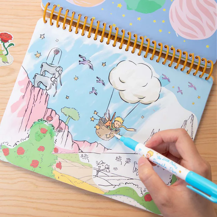 Livro de colorir do Pequeno Príncipe com caneta azul nas mãos sobre mesa de madeira