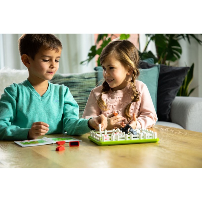 Crianças jogando jogo de tabuleiro com peças coloridas num tabuleiro verde com cercas brancas em sala com sofá e plantas.