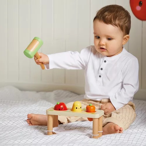 Criança a brincar com brinquedo de madeira e martelo coloridos sentada numa cama branca com padrão de quadrícula