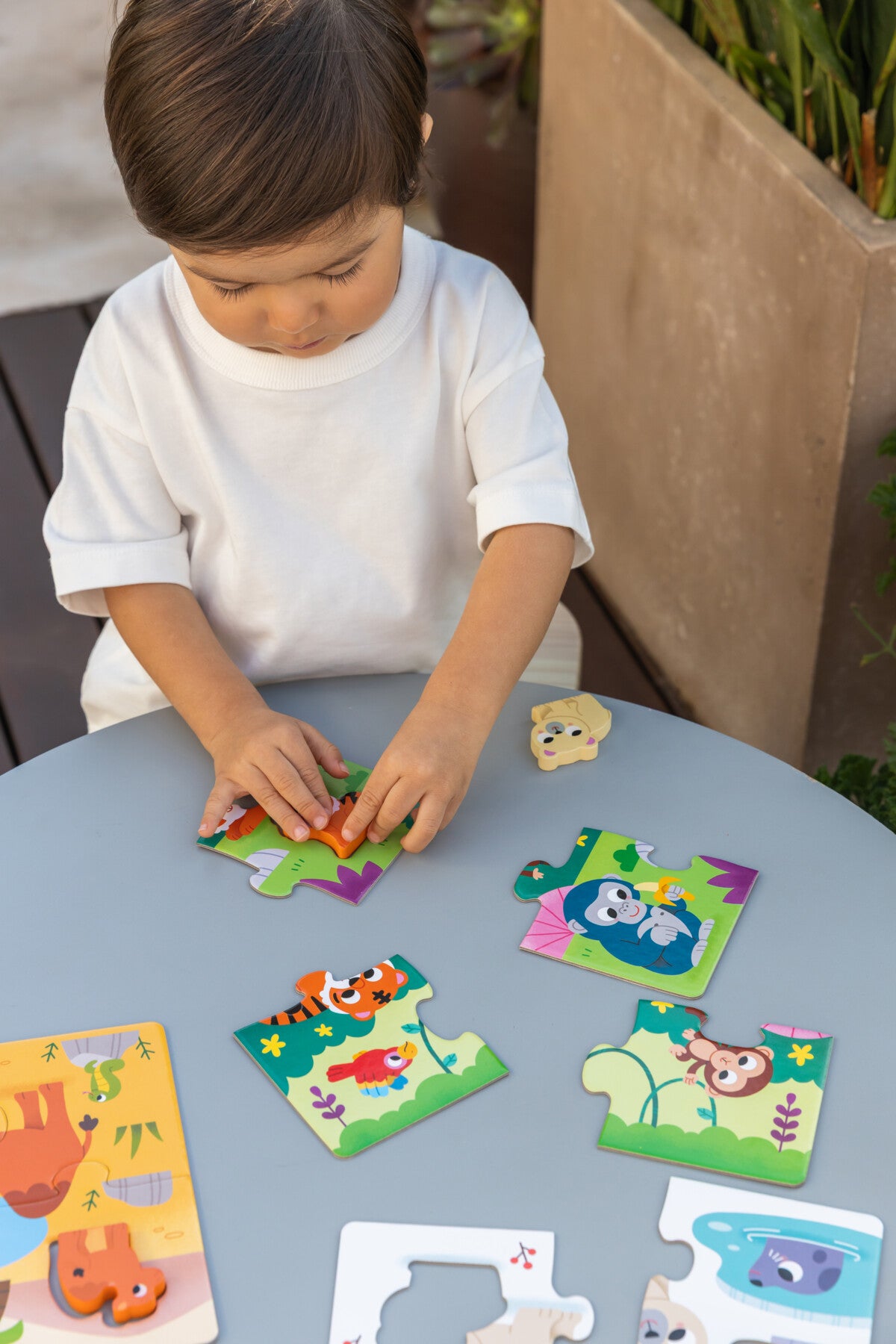 Criança montando puzzle de animais coloridos numa mesa cinzenta outdoors.