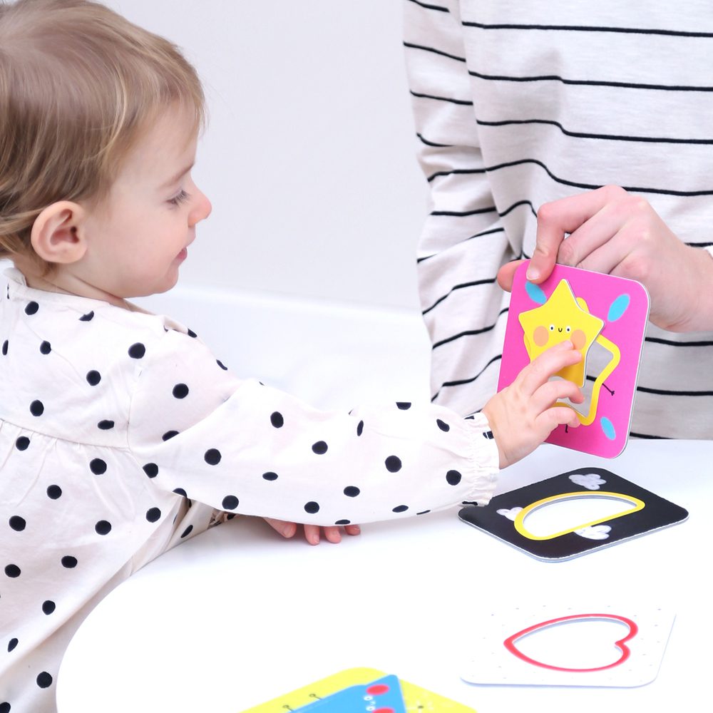 Criança pequena com camisola branca às bolinhas a brincar com adulto com camisola riscada, cartões coloridos numa mesa branca