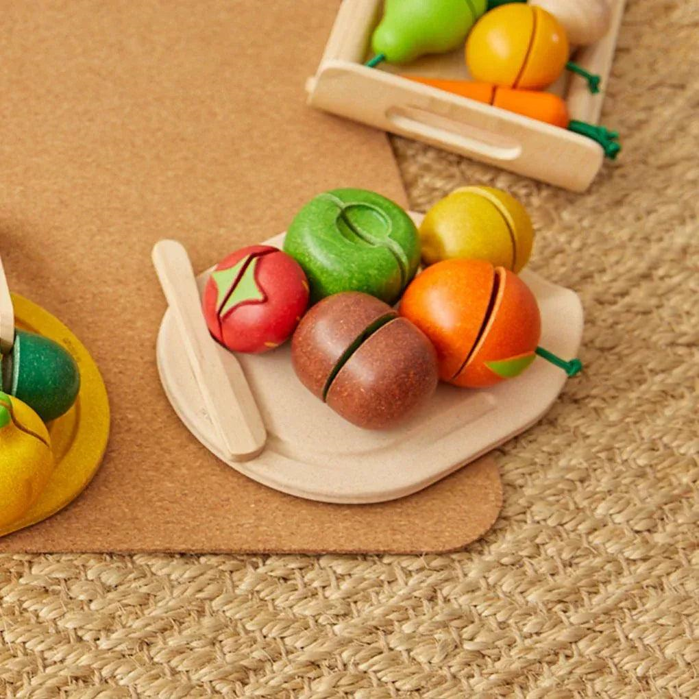 Brinquedos de madeira coloridos representando frutas com tábua e faca, sobre cortiça e tapete