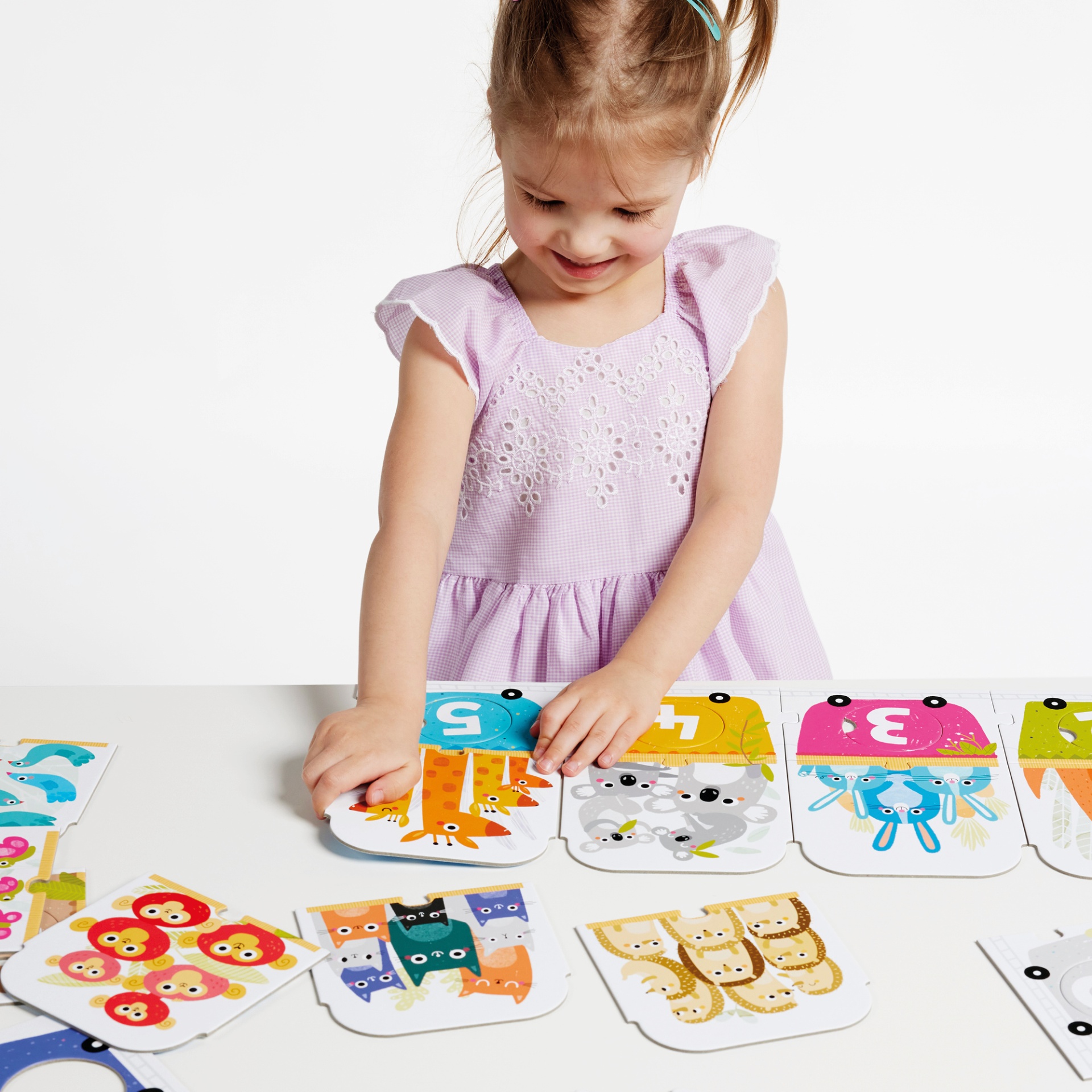 Criança com vestido lilás brincando com cartas educativas coloridas e numeradas com animais numa mesa branca.