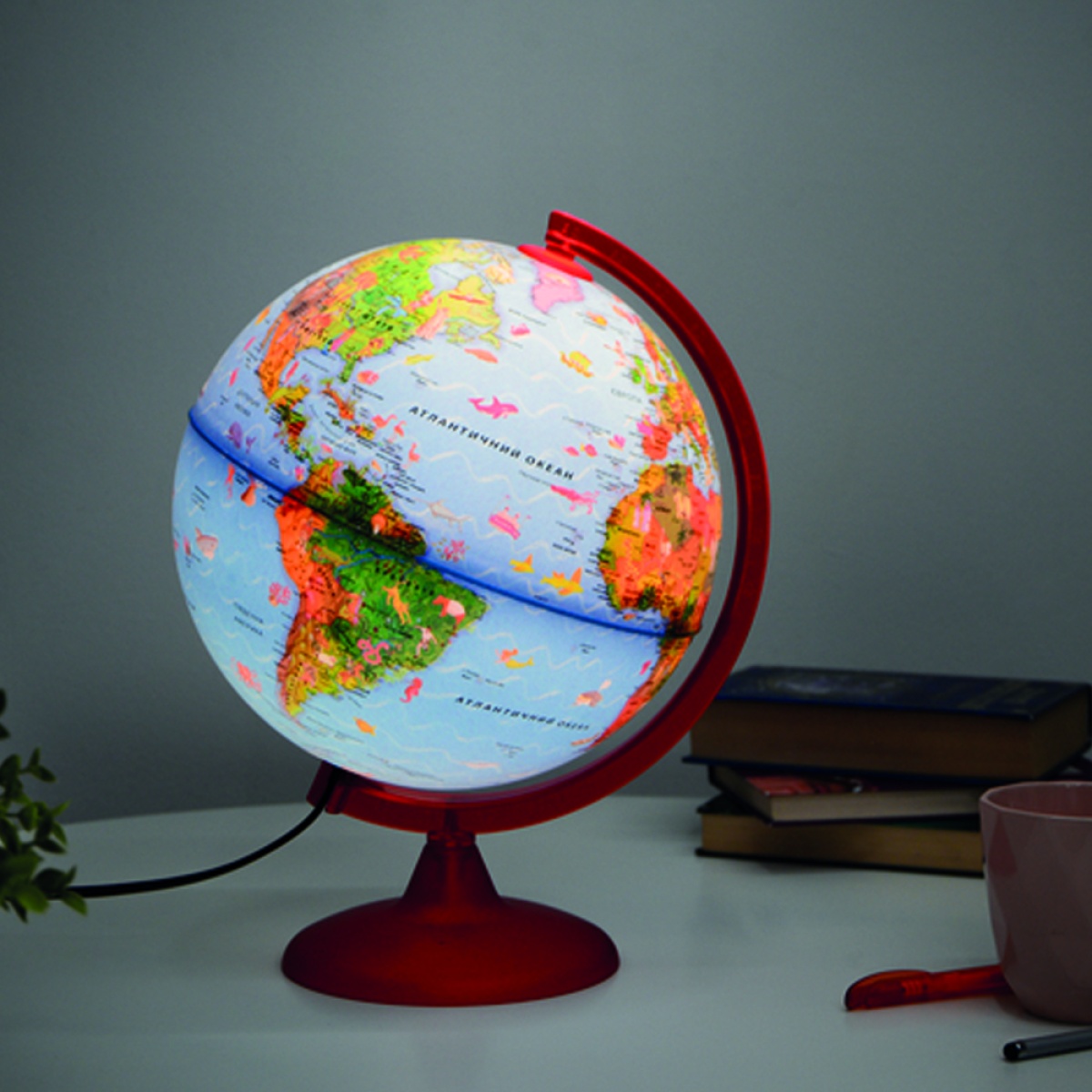 Globo terrestre luminoso com base vermelha numa mesa com livros, caneta e caneca.