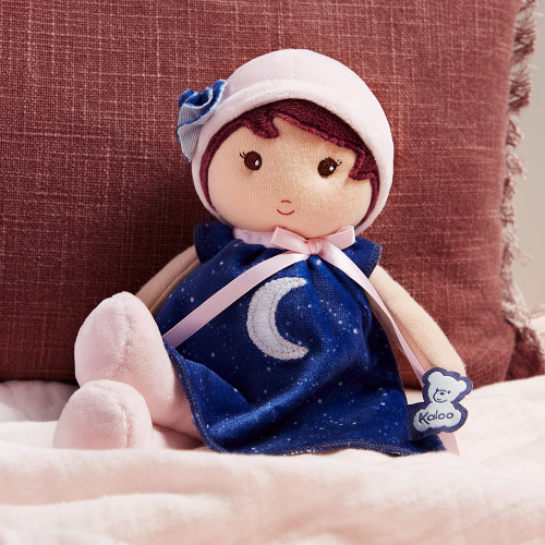 Boneca de peluche com vestido azul e chapéu rosa sentada numa cama cor-de-rosa