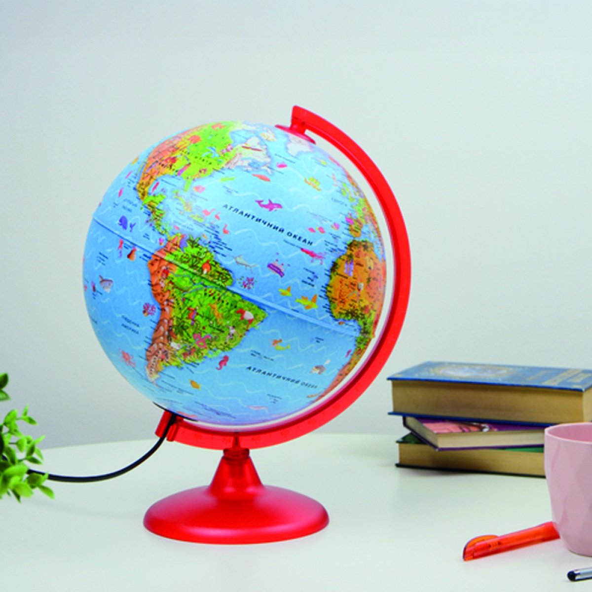 Globo terrestre colorido com suporte vermelho sobre mesa branca com livros, caneta e caneca.