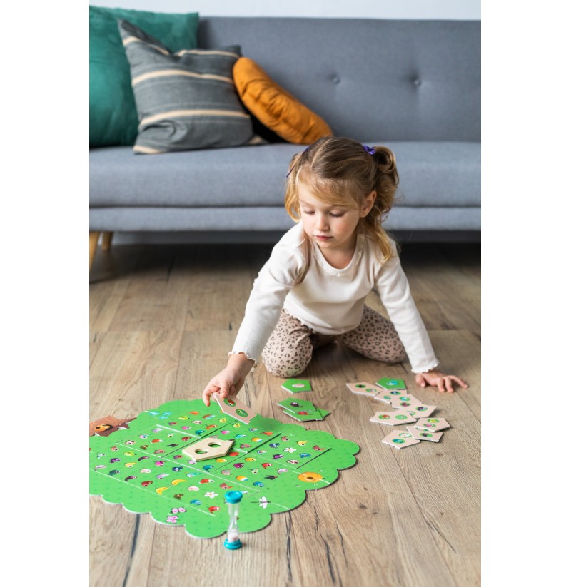 Menina a jogar jogo de tabuleiro verde em forma de árvore no chão de madeira com sofá e almofadas ao fundo.