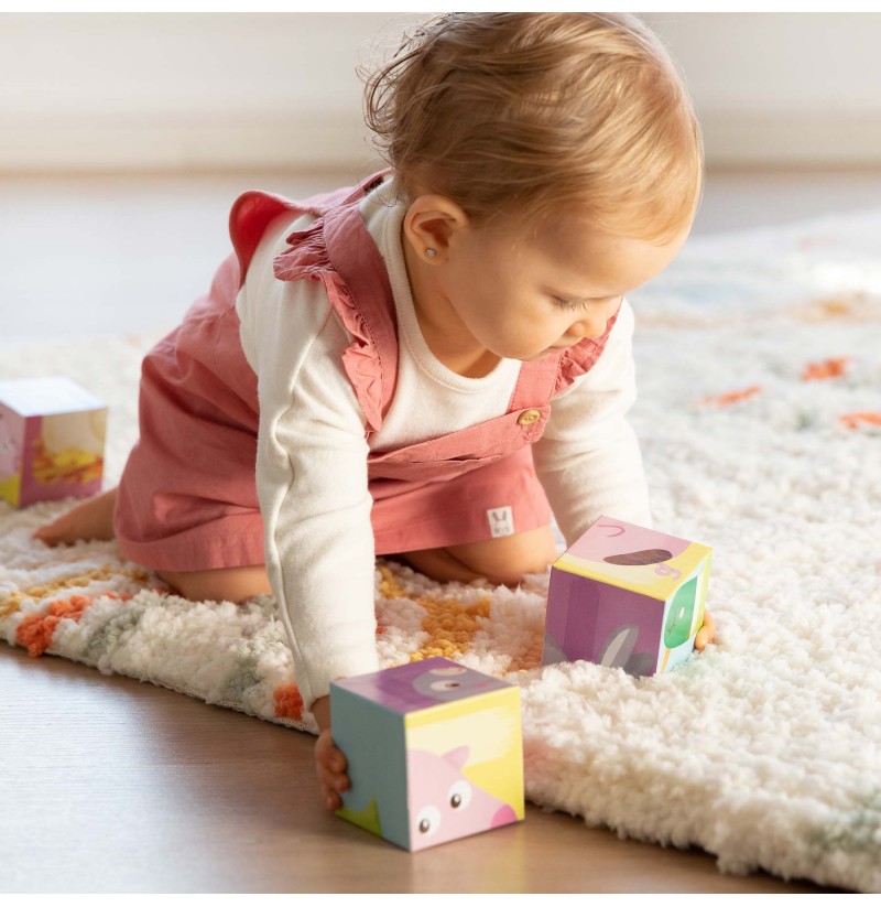 Bebé a brincar com blocos de madeira coloridos num tapete fofo branco com manchas cor-de-rosa e laranja