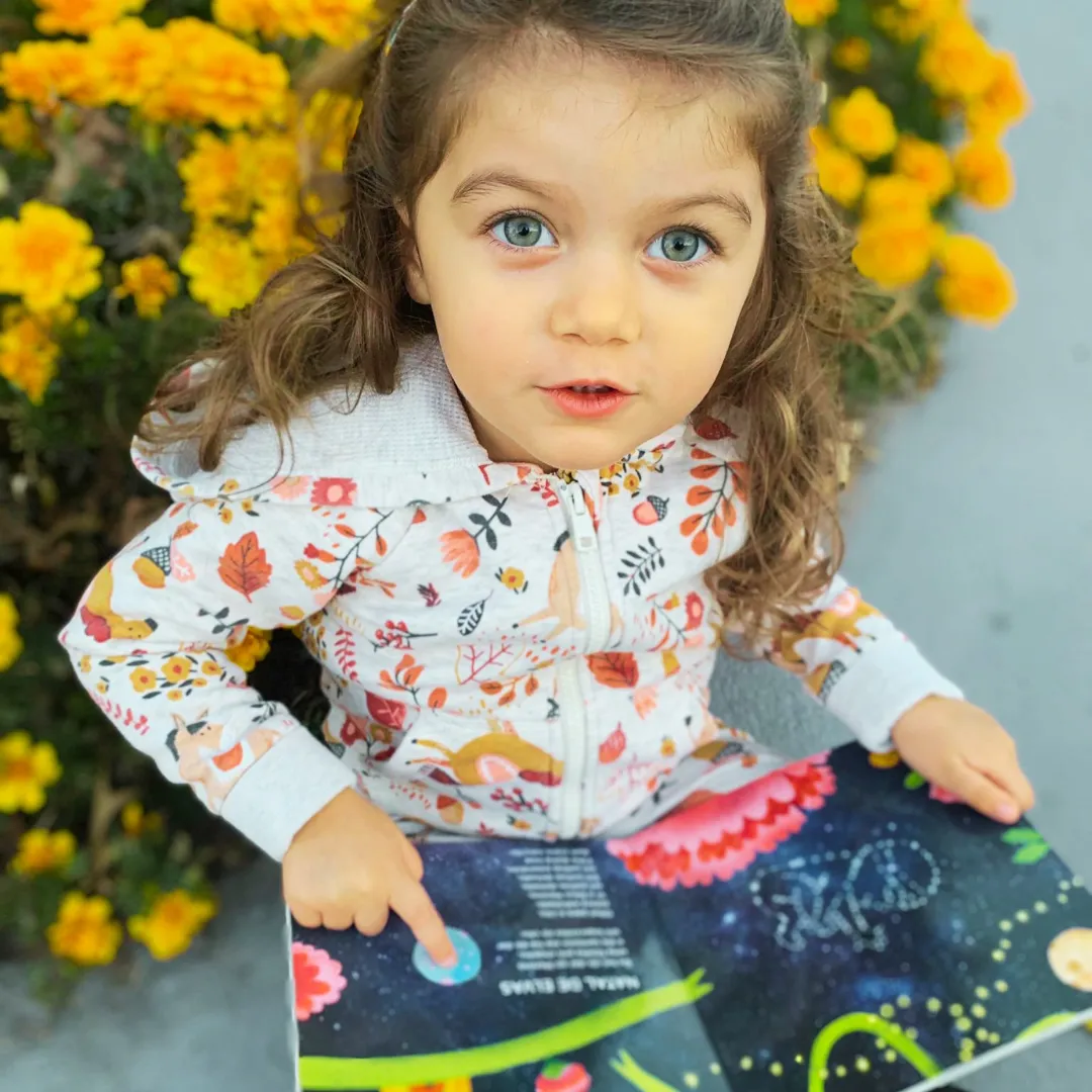 Menina com casaco estampado a apontar para um livro colorido sentada junto a flores amarelas