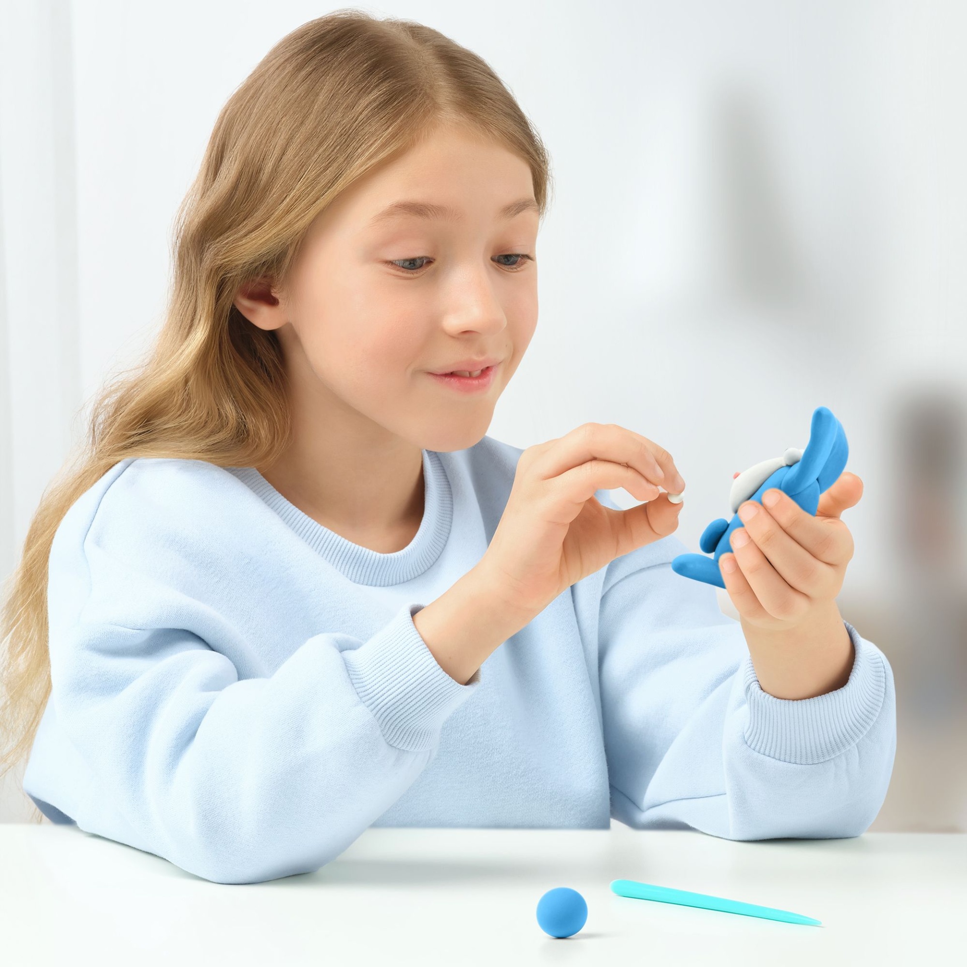 Criança a moldar plastilina azul e branca com as mãos sobre mesa branca.