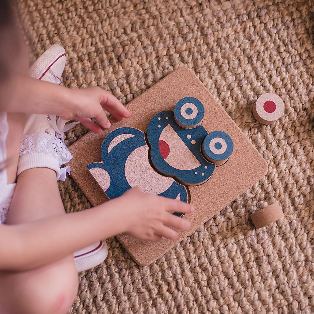 Brinquedo infantil de cortiça com figura azul e branca sobre base quadrada em tapete bege.