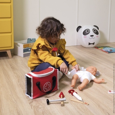 Criança a brincar com boneco bebê e acessórios médicos de brinquedo junto a um saco vermelho com cruz branca sobre chão de madeira clara