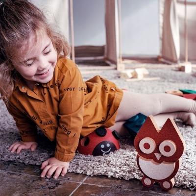 Criança a brincar com brinquedos de madeira em forma de coruja e joaninha num tapete cinza.