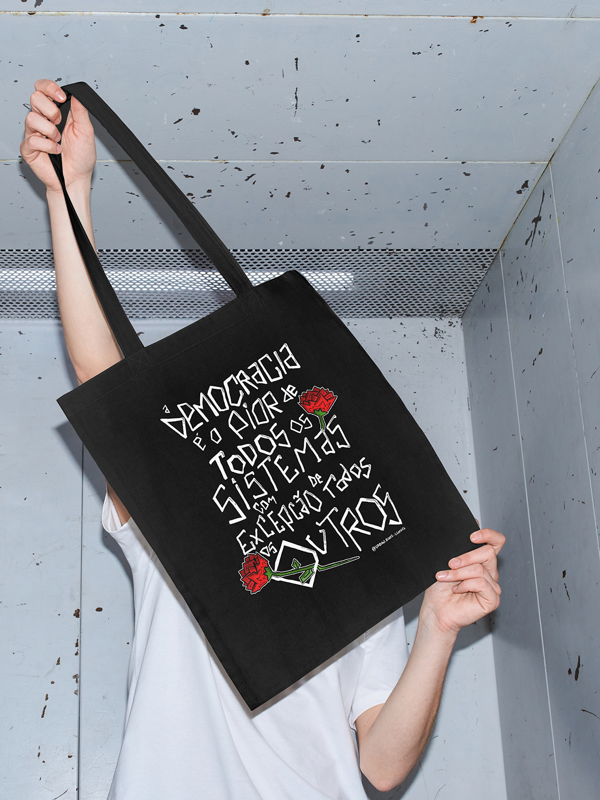 Mala de pano preta com texto branco e rosas vermelhas segurada por pessoa com t-shirt branca