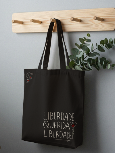 Saco de pano preto com texto e desenho de cravos pendurado em suporte de madeira
