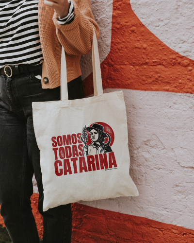 Saco de tecido com estampa vermelha e preta de mulher e texto em português, ao lado de parede texturada.