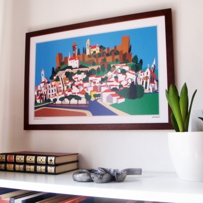 Pintura colorida de vila com castelo emoldurada na parede acima de estante branca com livros, vaso e objeto de pedra