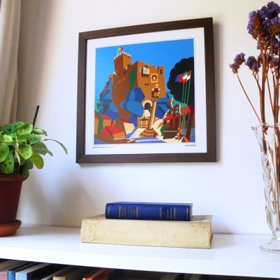 Quadro com arte de castelo colorido numa parede branca, com livros, planta em vaso e flores secas em superfície branca.