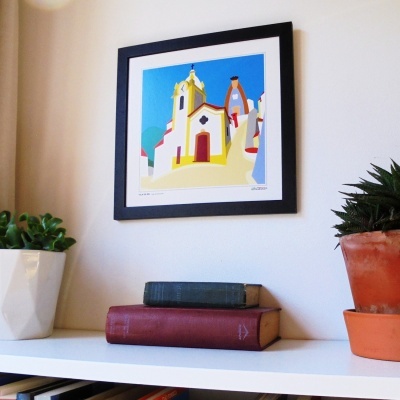 Impressão artística colorida de igreja emoldurada numa parede branca com plantas e livros numa prateleira