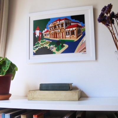 Gravura colorida de edifício antigo em moldura branca pendurada na parede com livros e plantas numa estante branca.