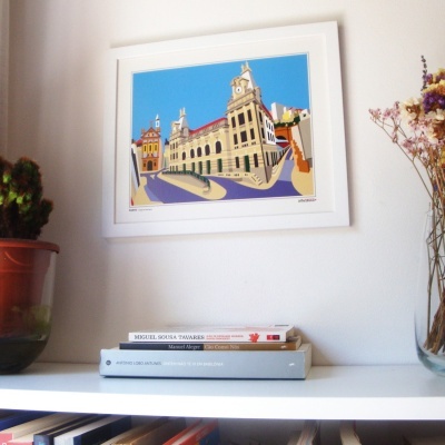 Impressão artística de edifício histórico colorido emoldurada na parede com livros e plantas na prateleira em madeira branca