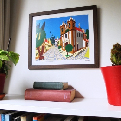 Quadro de pintura arquitetónica colorida com moldura castanha numa parede branca acima de estante branca com livros e plantas.