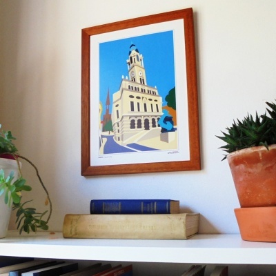 Quadro com ilustração de edifício em moldura de madeira na parede acima de prateleira com plantas e livros.