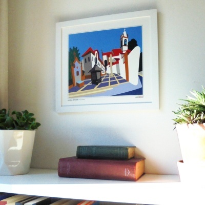 Quadro colorido de praça com moldura branca sobre estante branca com livros e plantas em vasos brancos