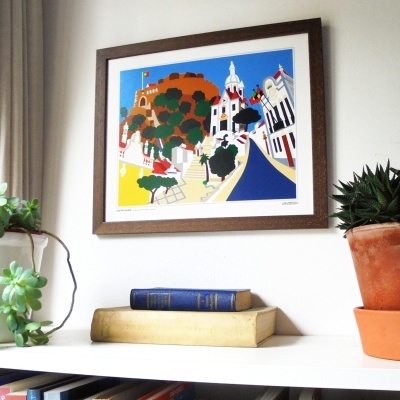 quadro colorido com paisagem urbana em moldura castanha, prateleira branca com livros e plantas