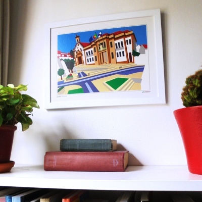 Quadro branco com pintura de edifícios coloridos e duas plantas com livros numa prateleira branca