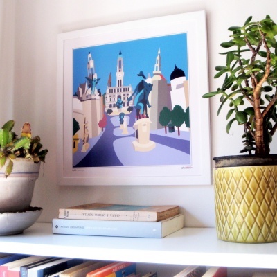 Ilustração urbana colorida em moldura branca na parede, com plantas em vasos e livros numa prateleira branca