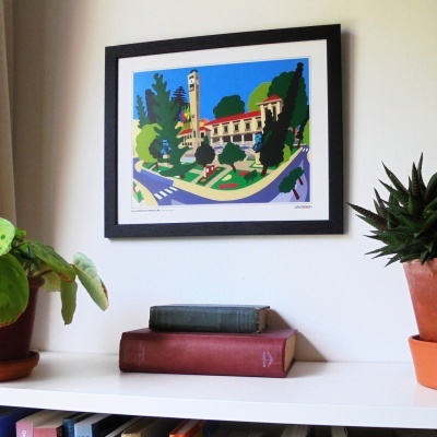 Quadro colorido de edifício em moldura preta na parede com livros e plantas na estante branca abaixo