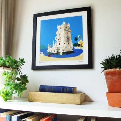 Pintura emoldurada da Torre de Belém na parede com plantas e livros numa mesa branca