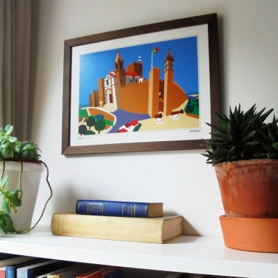 Pintura colorida emoldurada de castelo com plantas e livros numa prateleira
