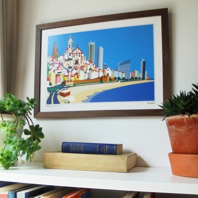 Pintura colorida de paisagem urbana costeira em moldura castanha, pendurada numa parede branca acima de uma estante com livros e plantas.