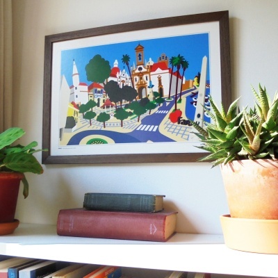 Pintura colorida de cidade em moldura pendurada numa parede acima de prateleira com livros e plantas