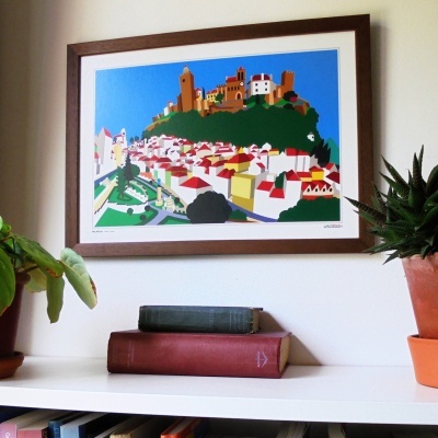 Quadro colorido de vila estilizada pendurado na parede, prateleira com livros e plantas