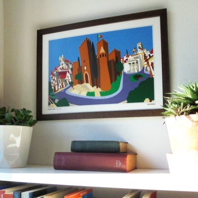 Quadro colorido com castelo e edifícios numa parede branca, acima de estante com livros e plantas.