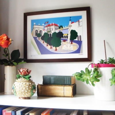 Estante branca com livros, vasos de plantas e uma pintura urbana colorida emoldurada na parede