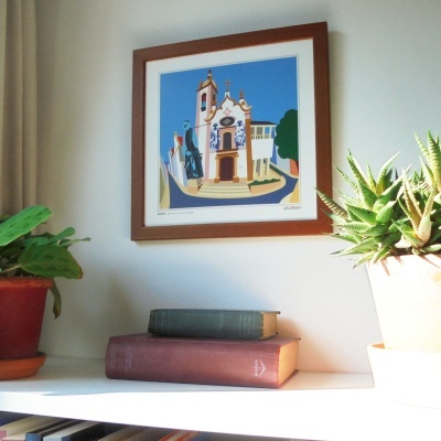 Estante com duas plantas em vasos, livros antigos e uma pintura de igreja emoldurada