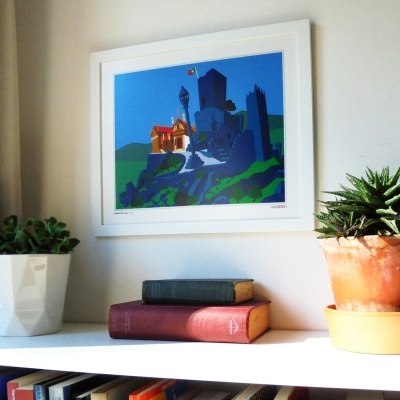 Quadro branco com gravura de castelo colorido pendurado numa parede, estante branca com livros e plantas em vasos