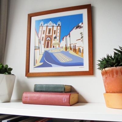 Impressão emoldurada de uma igreja com céu azul numa parede branca acima de estante com livros e plantas.