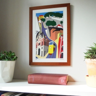 Quadro com ilustração colorida de bonde amarelo pendurado na parede acima de estante com livro e plantas