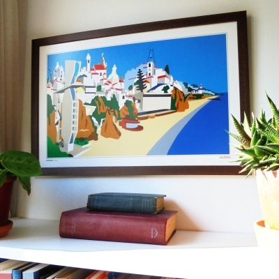 Quadro colorido de praia e edifícios em moldura preta, acima de prateleira com livros e plantas