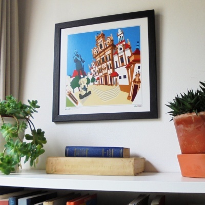 Quadro colorido de edifício antigo em moldura preta sobre prateleira com plantas e livros